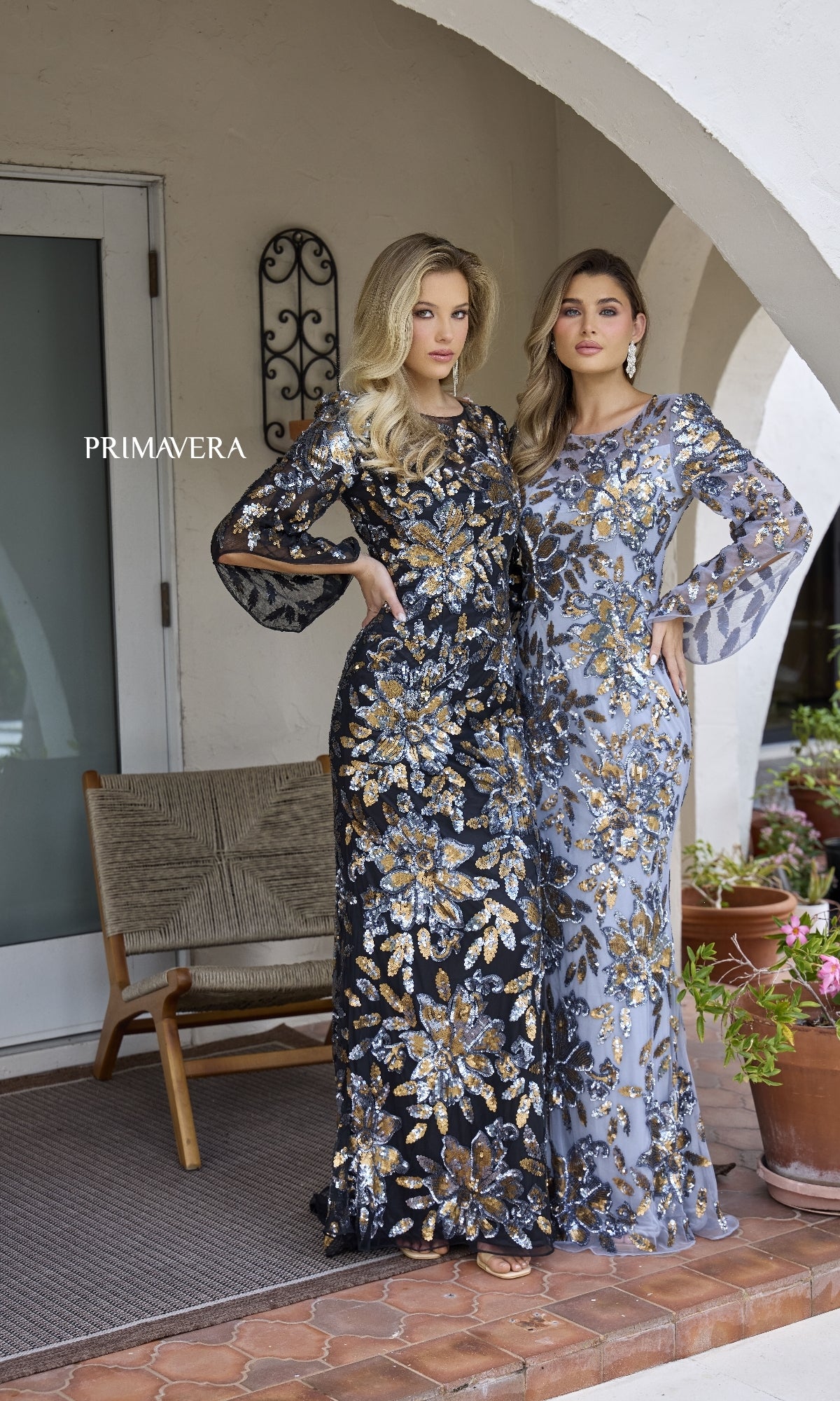 Two women wear Primavera 12171 long formal dresses—one in black and gold with sheer sleeves, the other in silver and gold—posing side by side on a tiled patio near a wooden bench and potted plants.